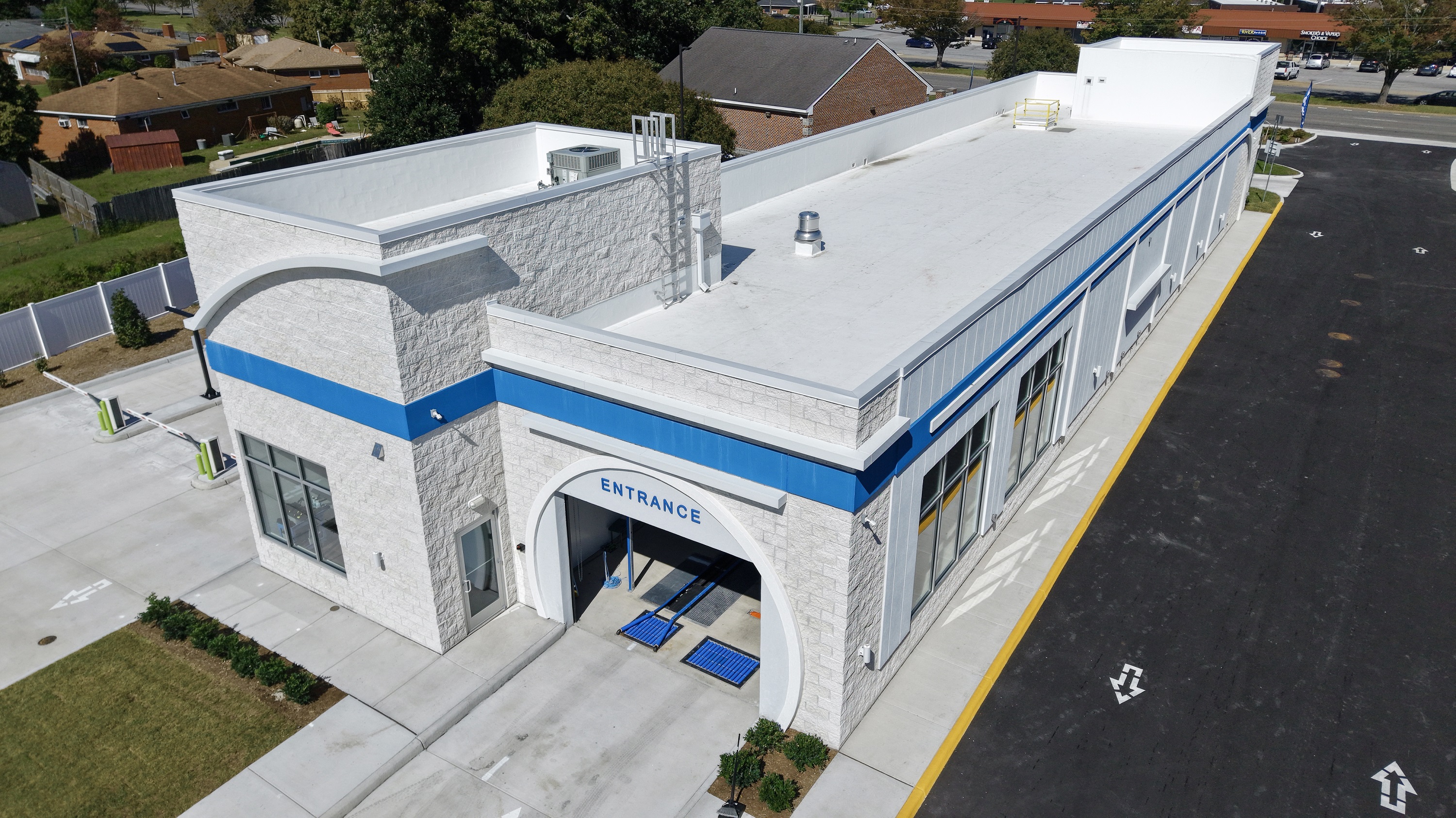 Aerial view of a newly constructed car wash building with a white exterior, blue accent stripe, and an arched entrance labeled “Entrance.” The structure features large windows along the side, a clean concrete driveway, and adjacent landscaped areas. Surrounding the site are residential homes, trees, and a nearby commercial area.