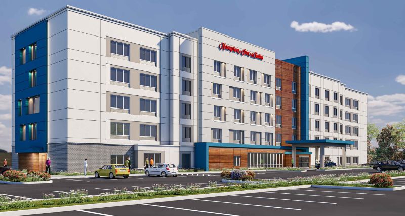 Architectural rendering of a five-story Hampton Inn by Hilton hotel featuring a modern design with white and gray exterior panels, blue accent sections, and wood-tone siding. Large windows line the façade, with a covered entrance canopy at the front. The surrounding area includes landscaped plantings, a parking lot, and a clear sky with scattered clouds.