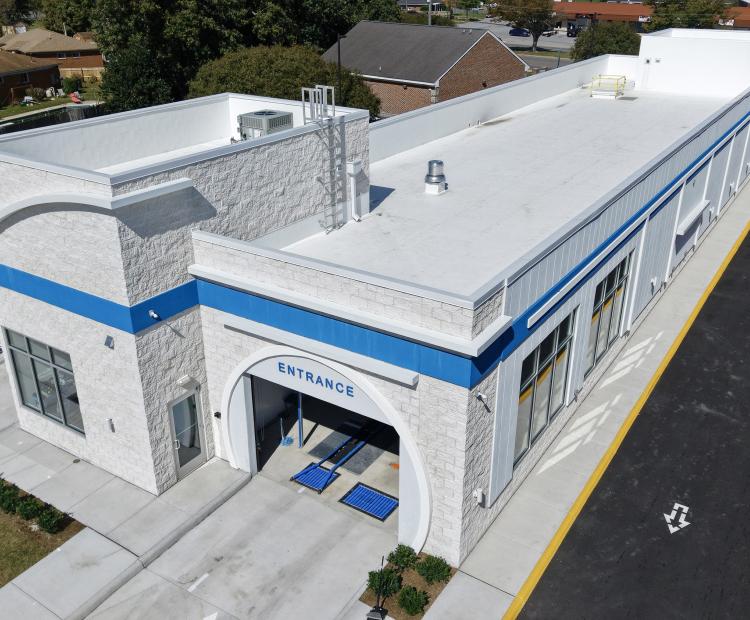 Aerial view of a newly constructed car wash building with a white exterior, blue accent stripe, and an arched entrance labeled “Entrance.” The structure features large windows along the side, a clean concrete driveway, and adjacent landscaped areas. Surrounding the site are residential homes, trees, and a nearby commercial area.