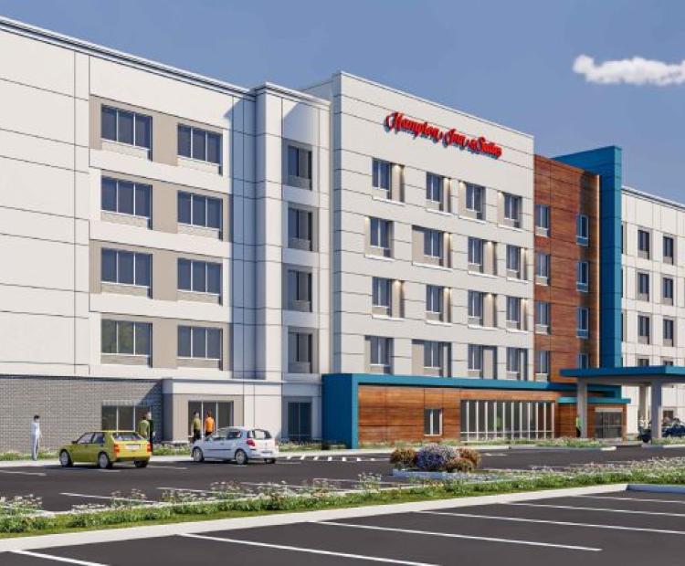 Architectural rendering of a five-story Hampton Inn by Hilton hotel featuring a modern design with white and gray exterior panels, blue accent sections, and wood-tone siding. Large windows line the façade, with a covered entrance canopy at the front. The surrounding area includes landscaped plantings, a parking lot, and a clear sky with scattered clouds.