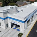 Aerial view of a newly constructed car wash building with a white exterior, blue accent stripe, and an arched entrance labeled “Entrance.” The structure features large windows along the side, a clean concrete driveway, and adjacent landscaped areas. Surrounding the site are residential homes, trees, and a nearby commercial area.