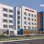 Architectural rendering of a five-story Hampton Inn by Hilton hotel featuring a modern design with white and gray exterior panels, blue accent sections, and wood-tone siding. Large windows line the façade, with a covered entrance canopy at the front. The surrounding area includes landscaped plantings, a parking lot, and a clear sky with scattered clouds.